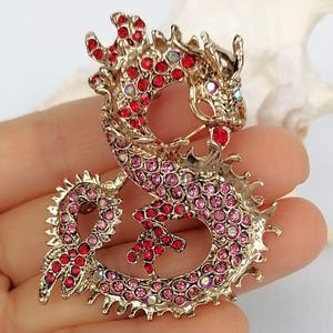 Puede incluir: Broche de drag&oacute;n dorado, incrustado con pedrer&iacute;a roja y rosa. El drag&oacute;n est&aacute; en una pose curvada, con escamas detalladas y un cierre de alfiler. Un accesorio decorativo.