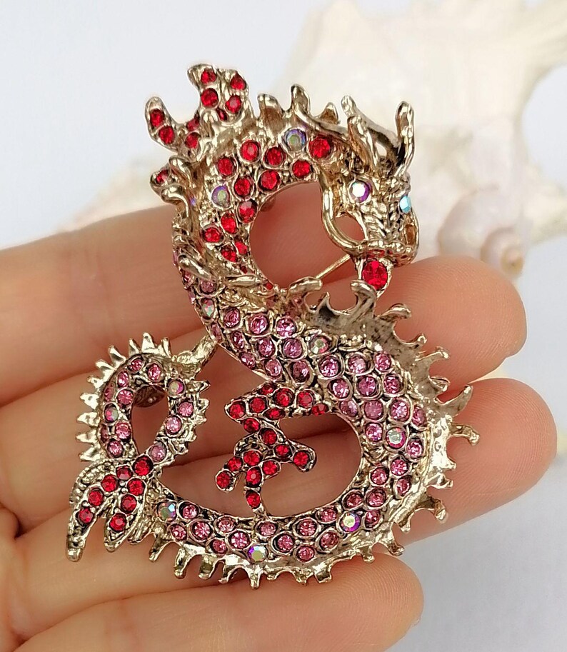Puede incluir: Broche de drag&oacute;n dorado, incrustado con pedrer&iacute;a roja y rosa. El drag&oacute;n est&aacute; en una pose curvada, con escamas detalladas y un cierre de alfiler. Un accesorio decorativo.