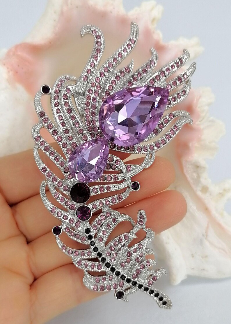 May include: A silver-toned feather brooch featuring two large, pear-shaped lavender crystals, accented with smaller round pink crystals and a row of dark purple stones along the central spine, creating a detailed and textured metallic design.