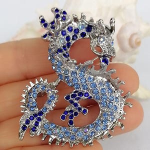 Puede incluir: Broche de drag&oacute;n plateado, incrustado con pedrer&iacute;a azul de diferentes tonos. El drag&oacute;n est&aacute; en una pose curvada, con escamas detalladas y una expresi&oacute;n feroz. Es una joya.