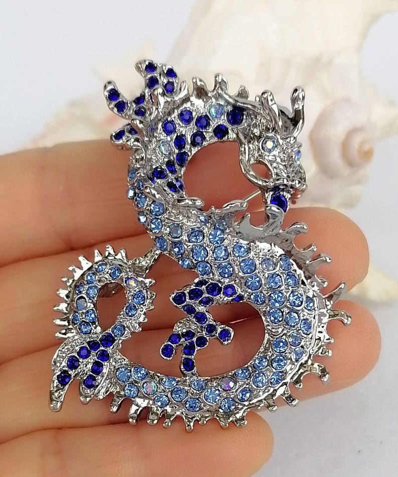 Puede incluir: Broche de drag&oacute;n plateado, incrustado con pedrer&iacute;a azul de diferentes tonos. El drag&oacute;n est&aacute; en una pose curvada, con escamas detalladas y una expresi&oacute;n feroz. Es una joya.