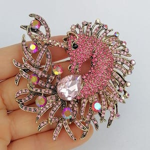 May include: A pink and gold-toned unicorn brooch. The unicorn is encrusted with pink rhinestones, with a large teardrop-shaped pink gem. The brooch has a gold-toned metal frame with pink rhinestone accents and iridescent gems.