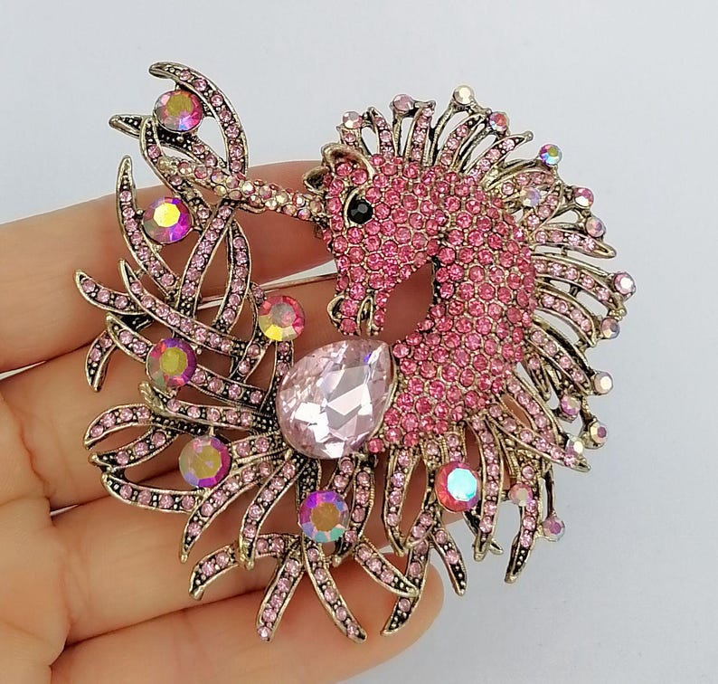 May include: A pink and gold-toned unicorn brooch. The unicorn is encrusted with pink rhinestones, with a large teardrop-shaped pink gem. The brooch has a gold-toned metal frame with pink rhinestone accents and iridescent gems.