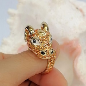 May include: A gold-toned ring shaped like a bull's head, featuring a polished metal finish on the horns and ears, with the face and band encrusted in small, sparkling orange crystals and accented with white and black enamel around the eyes.