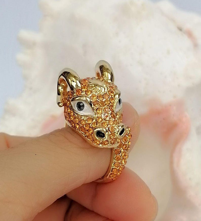 May include: A gold-toned ring shaped like a bull's head, featuring a polished metal finish on the horns and ears, with the face and band encrusted in small, sparkling orange crystals and accented with white and black enamel around the eyes.
