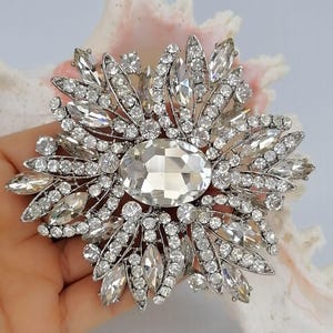 Op de afbeelding: Een zilverkleurige broche met een grote centrale ovale heldere kristal, omringd door een zonnestraalpatroon van kleinere ronde en marquise-geslepen heldere kristallen. De metalen zetting is gedetailleerd en houdt talloze fonkelende stenen vast.