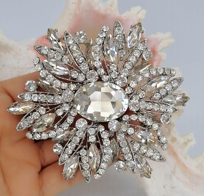 Op de afbeelding: Een zilverkleurige broche met een grote centrale ovale heldere kristal, omringd door een zonnestraalpatroon van kleinere ronde en marquise-geslepen heldere kristallen. De metalen zetting is gedetailleerd en houdt talloze fonkelende stenen vast.