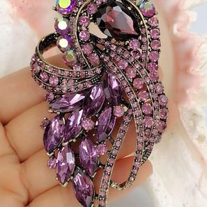 May include: A decorative brooch featuring a dark purple pear-shaped center stone surrounded by smaller light purple round crystals and deep purple marquise-cut stones. The metal setting is an antique bronze tone with a curved, flowing design.