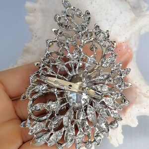 May include: A silver-toned metal brooch featuring an intricate, openwork filigree design shaped like a teardrop. The piece is encrusted with numerous clear, faceted crystals and includes a horizontal pin mechanism attached to the back.