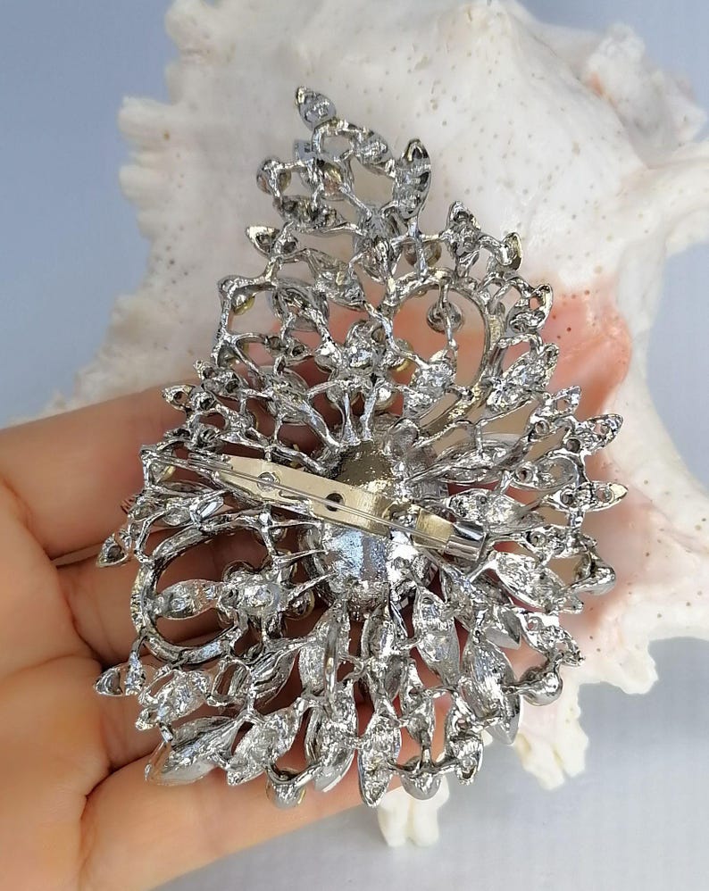 May include: A silver-toned metal brooch featuring an intricate, openwork filigree design shaped like a teardrop. The piece is encrusted with numerous clear, faceted crystals and includes a horizontal pin mechanism attached to the back.