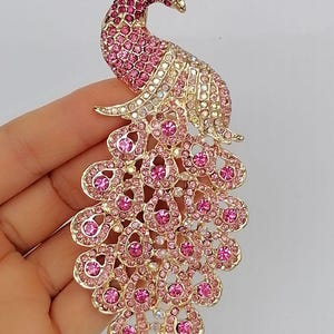 May include: A gold-toned peacock brooch with pink and clear crystals. The brooch features a detailed design with a fanned tail, a curved neck, and a small crown. The crystals are arranged to create a sparkling effect.