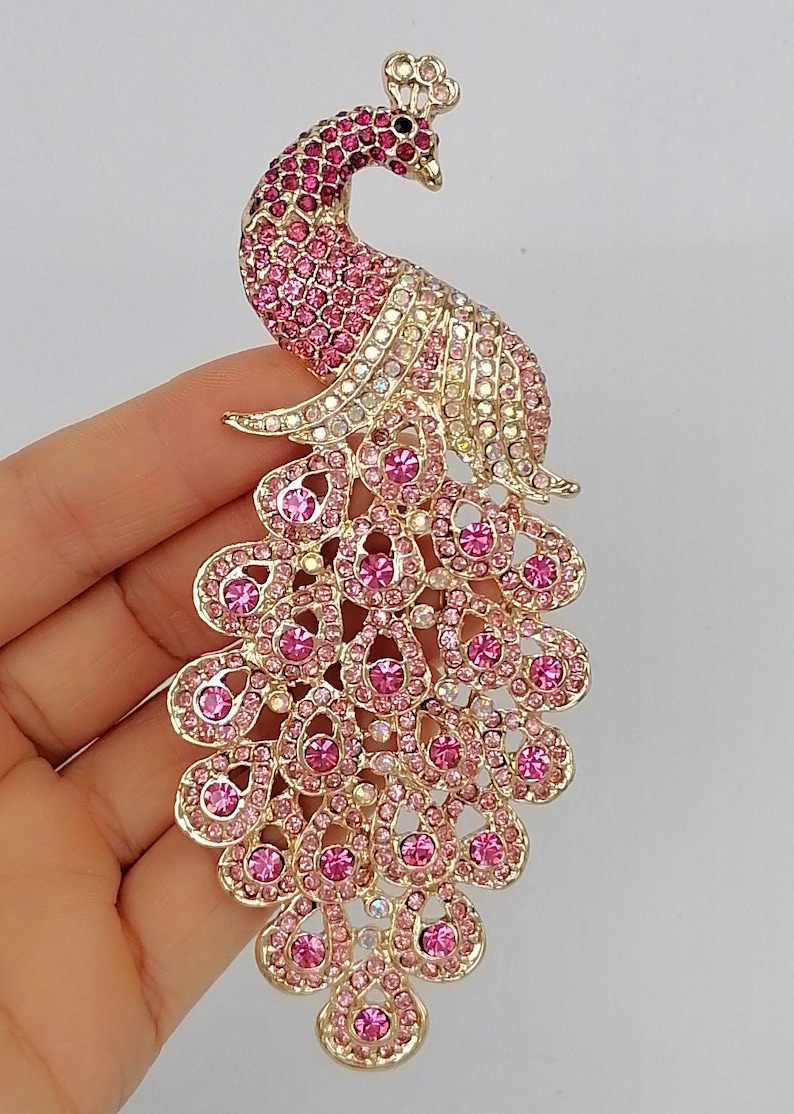 May include: A gold-toned peacock brooch with pink and clear crystals. The brooch features a detailed design with a fanned tail, a curved neck, and a small crown. The crystals are arranged to create a sparkling effect.