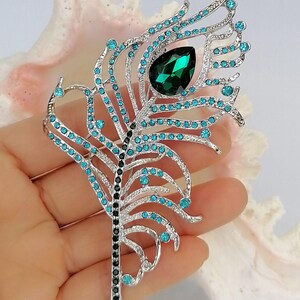 May include: A silver-coloured brooch in the shape of a peacock feather, decorated with teal and emerald green gemstones. The brooch has a large, teardrop-shaped green stone at the centre and is accented with numerous smaller teal stones.