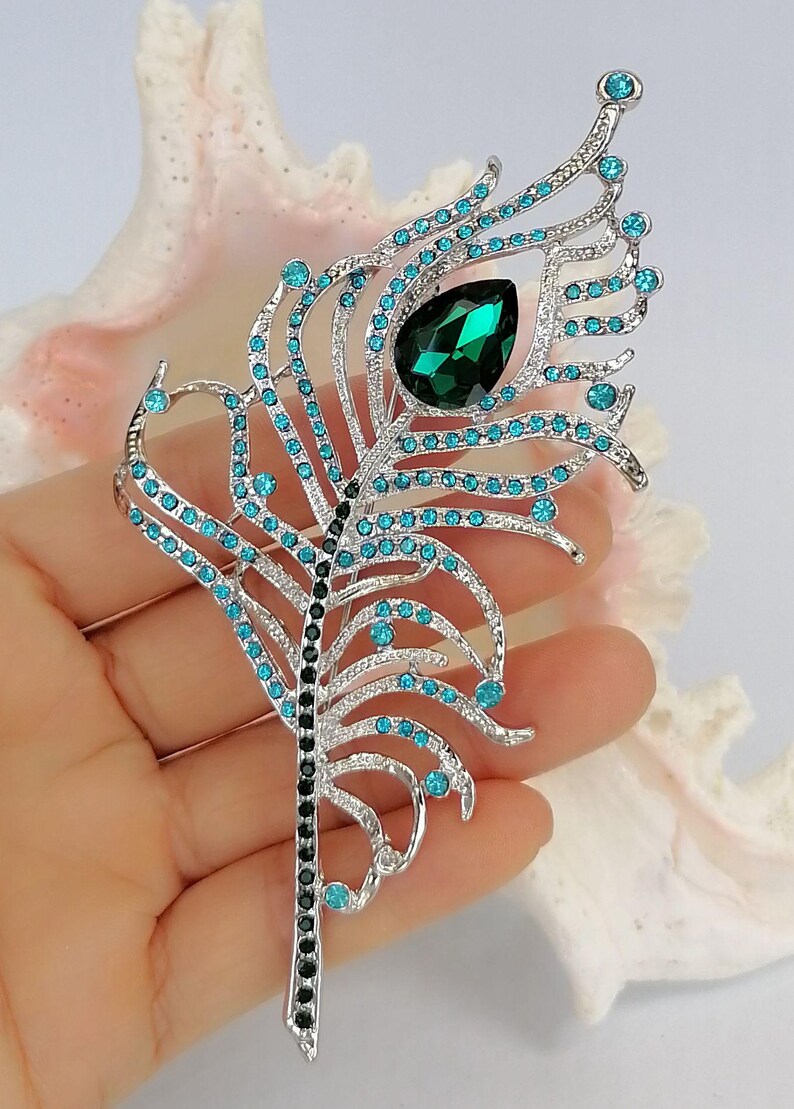 May include: A silver-coloured brooch in the shape of a peacock feather, decorated with teal and emerald green gemstones. The brooch has a large, teardrop-shaped green stone at the centre and is accented with numerous smaller teal stones.