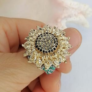 K&ouml;nnte beinhalten: Ein sonnenblumenf&ouml;rmiger Ring mit goldfarbenen Bl&uuml;tenbl&auml;ttern, einem Zentrum aus dunkelgrauen und schwarzen Steinen sowie kleinen gr&uuml;nen Steinen an der Basis der Blume. Das Design ist detailreich mit geschichteten Bl&uuml;tenbl&auml;ttern und metallischer Oberfl&auml;che.
