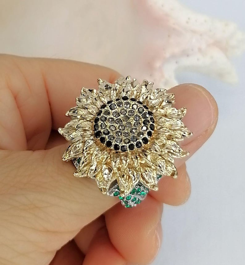 K&ouml;nnte beinhalten: Ein sonnenblumenf&ouml;rmiger Ring mit goldfarbenen Bl&uuml;tenbl&auml;ttern, einem Zentrum aus dunkelgrauen und schwarzen Steinen sowie kleinen gr&uuml;nen Steinen an der Basis der Blume. Das Design ist detailreich mit geschichteten Bl&uuml;tenbl&auml;ttern und metallischer Oberfl&auml;che.
