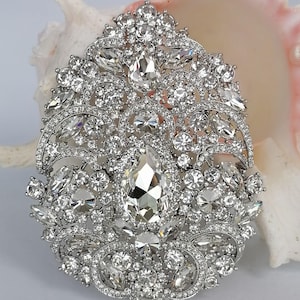 May include: A silver-toned brooch with a dazzling array of clear, sparkling crystals. The brooch features a large, pear-shaped central stone surrounded by smaller, round and marquise-cut crystals, creating an ornate, elegant design. The brooch is set against a soft, neutral background.
