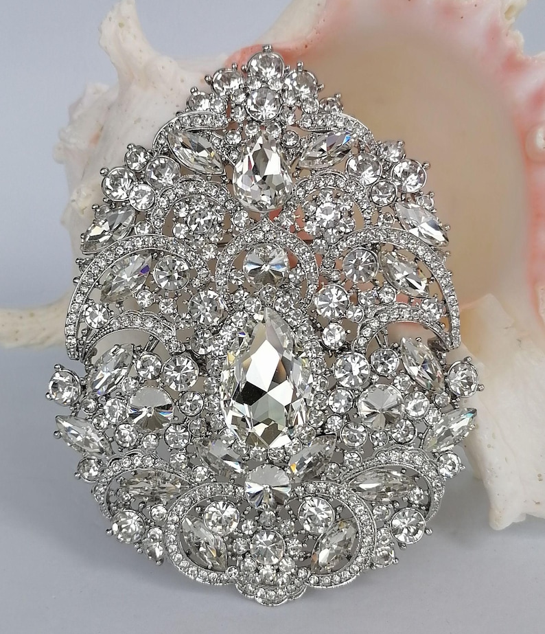 May include: A silver-toned brooch with a dazzling array of clear, sparkling crystals. The brooch features a large, pear-shaped central stone surrounded by smaller, round and marquise-cut crystals, creating an ornate, elegant design. The brooch is set against a soft, neutral background.