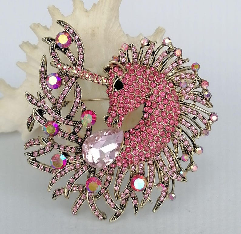May include: A pink unicorn brooch with a gold-tone metal frame. The unicorn is encrusted with pink rhinestones and has a large, pear-shaped pink crystal. Iridescent crystals and decorative leaves complete the design. A pin is attached to the back.