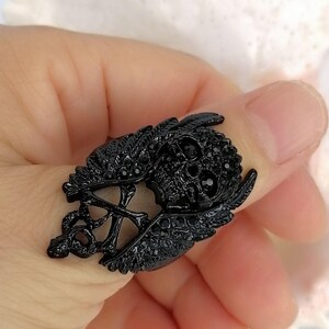 以下が含まれることがあります： 小さな黒いクリスタルで装飾された中央の頭蓋骨モチーフが特徴の黒い指輪。頭蓋骨の両側には詳細な翼があり、交差した骨のペアの上に配置されています。全体がダークなモノクロの黒いトーンで仕上げられています。