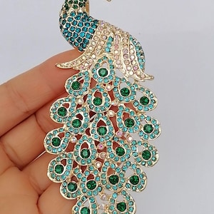 May include: A gold-toned peacock brooch with a detailed design. The peacock features a head and neck in shades of green and teal, with a body and tail adorned with teal and green gemstones. The brooch is an eye-catching accessory.