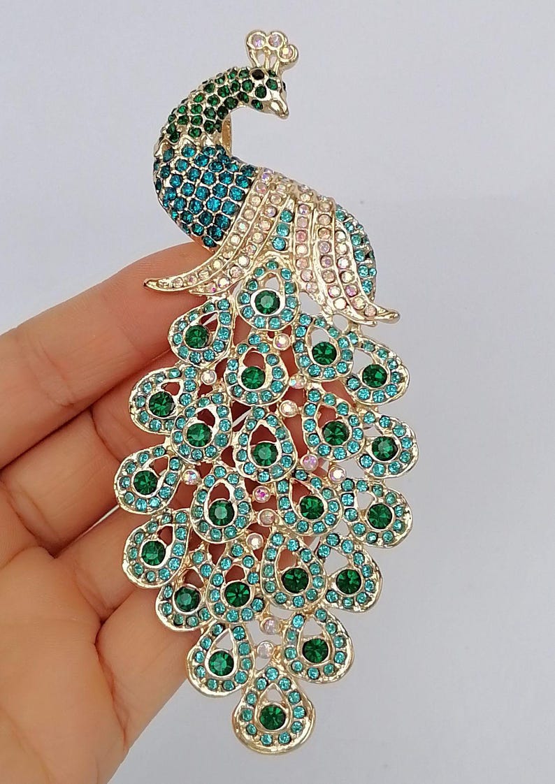 May include: A gold-toned peacock brooch with a detailed design. The peacock features a head and neck in shades of green and teal, with a body and tail adorned with teal and green gemstones. The brooch is an eye-catching accessory.