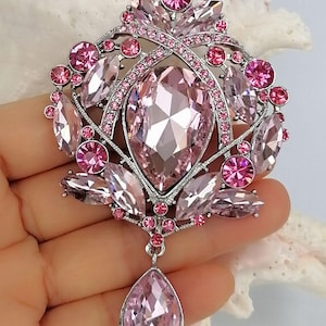 May include: A pink and silver brooch with a large, oval-shaped central stone and smaller, surrounding pink gems. The brooch has a teardrop-shaped pendant and a silver-toned metal setting. The design is ornate and features various shades of pink.