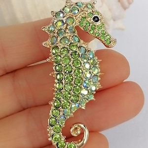 May include: A gold-toned seahorse brooch encrusted with green and iridescent crystals. The seahorse has a detailed design with a curled tail and a black eye. The brooch is a decorative accessory.