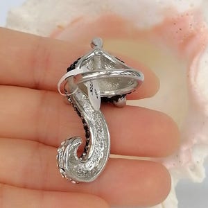 K&ouml;nnte beinhalten: Ein silberfarbener Ring mit einem geschwungenen, strukturierten Design, das mit kleinen schwarzen Kristallen verziert ist. Am Ring befindet sich ein wei&szlig;es Etikett mit der deutlich lesbaren Aufschrift 'SIZE 8', die die Ringgr&ouml;&szlig;e angibt.