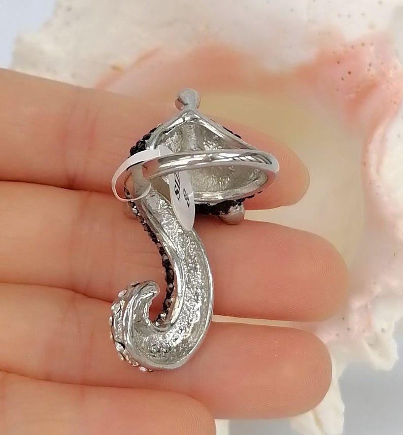 K&ouml;nnte beinhalten: Ein silberfarbener Ring mit einem geschwungenen, strukturierten Design, das mit kleinen schwarzen Kristallen verziert ist. Am Ring befindet sich ein wei&szlig;es Etikett mit der deutlich lesbaren Aufschrift 'SIZE 8', die die Ringgr&ouml;&szlig;e angibt.