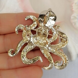 K&ouml;nnte beinhalten: Goldfarbene Oktopus-Brosche mit aufwendigen Details. Die Brosche hat acht sich kr&auml;uselnde Tentakel und eine strukturierte Oberfl&auml;che. Die Anstecknadel ist zum Tragen an Kleidung oder Accessoires konzipiert und verleiht einen maritimen Touch.