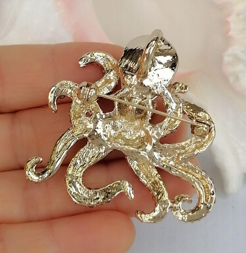 K&ouml;nnte beinhalten: Goldfarbene Oktopus-Brosche mit aufwendigen Details. Die Brosche hat acht sich kr&auml;uselnde Tentakel und eine strukturierte Oberfl&auml;che. Die Anstecknadel ist zum Tragen an Kleidung oder Accessoires konzipiert und verleiht einen maritimen Touch.