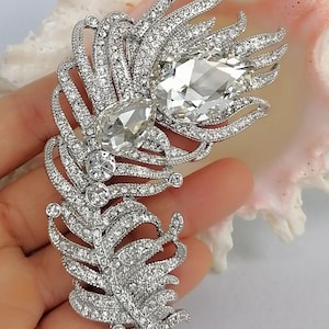 May include: A silver-toned feather brooch encrusted with numerous small clear rhinestones. The design features two prominent, large, pear-shaped clear crystals set within the upper section of the feather structure, creating a focal point.