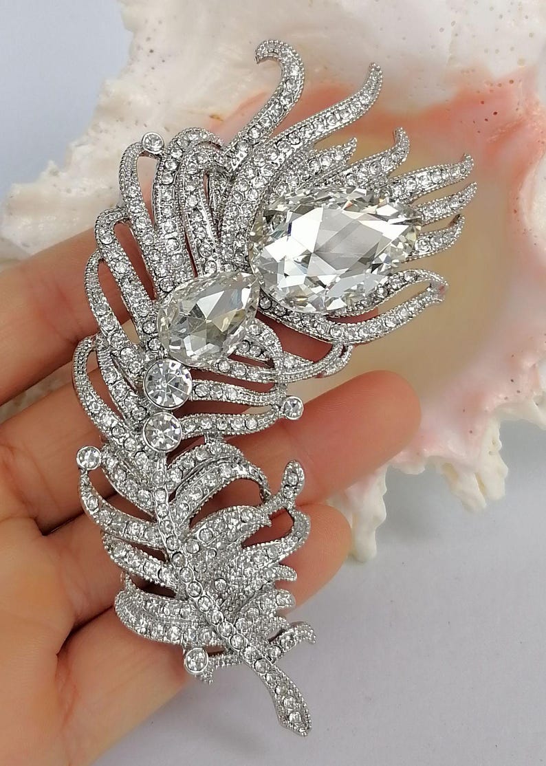 May include: A silver-toned feather brooch encrusted with numerous small clear rhinestones. The design features two prominent, large, pear-shaped clear crystals set within the upper section of the feather structure, creating a focal point.