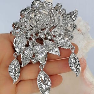 Puede incluir: Un broche de metal plateado con un dise&ntilde;o floral detallado de p&eacute;talos y hojas texturizadas. La parte posterior del broche incluye un mecanismo de alfiler horizontal y tres dijes colgantes en forma de hoja sujetos a la estructura.