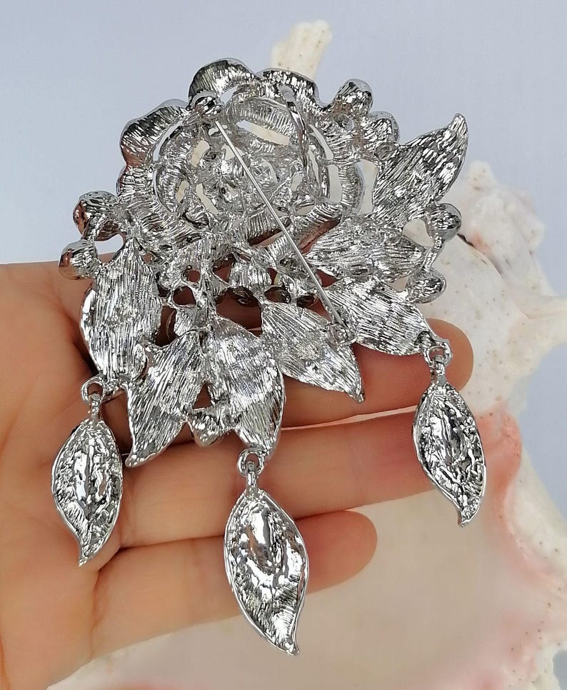 Puede incluir: Un broche de metal plateado con un dise&ntilde;o floral detallado de p&eacute;talos y hojas texturizadas. La parte posterior del broche incluye un mecanismo de alfiler horizontal y tres dijes colgantes en forma de hoja sujetos a la estructura.