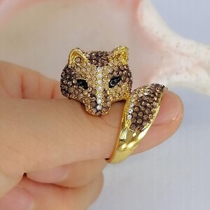 Pu&ograve; includere: Un anello color oro con un design a testa di volpe incastonato con cristalli marroni, beige e trasparenti. La testa di volpe &egrave; posizionata in alto, con una fascia curva a forma di coda che scende verso il basso, anch'essa decorata con cristalli.