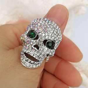 May include: A silver-toned ring shaped like a human skull, entirely encrusted with small, iridescent clear crystals. The skull features two prominent, round, deep green faceted stones set within the eye sockets, creating a striking contrast.