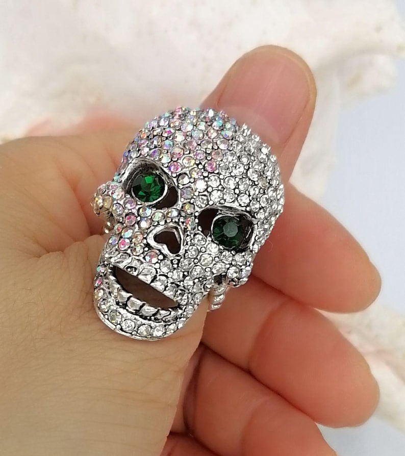 May include: A silver-toned ring shaped like a human skull, entirely encrusted with small, iridescent clear crystals. The skull features two prominent, round, deep green faceted stones set within the eye sockets, creating a striking contrast.