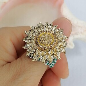 K&ouml;nnte beinhalten: Ein sonnenblumenf&ouml;rmiger Ring mit goldfarbenem Metallfinish und einem Zentrum aus gelben und braunen Kristallen. Die Bl&uuml;tenbl&auml;tter sind strukturiert und detailliert, und die Ringschiene ist mit kleinen, hellgr&uuml;nen Kristallen besetzt.