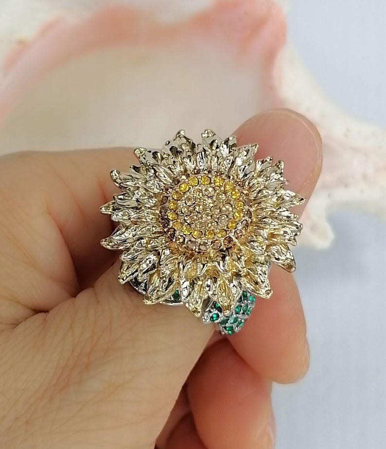 K&ouml;nnte beinhalten: Ein sonnenblumenf&ouml;rmiger Ring mit goldfarbenem Metallfinish und einem Zentrum aus gelben und braunen Kristallen. Die Bl&uuml;tenbl&auml;tter sind strukturiert und detailliert, und die Ringschiene ist mit kleinen, hellgr&uuml;nen Kristallen besetzt.