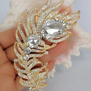 May include: A gold-toned feather-shaped brooch featuring a textured metal finish, encrusted with numerous small clear rhinestones, and accented with two large, prominent, pear-shaped clear crystals positioned along the central spine.