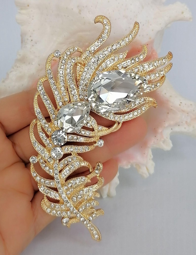 May include: A gold-toned feather-shaped brooch featuring a textured metal finish, encrusted with numerous small clear rhinestones, and accented with two large, prominent, pear-shaped clear crystals positioned along the central spine.