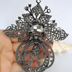 May include: A large, ornate brooch in a dark gunmetal finish, featuring a central rectangular gray crystal, surrounded by intricate filigree metalwork and numerous small, sparkling gray rhinestones arranged in floral and scroll patterns.
