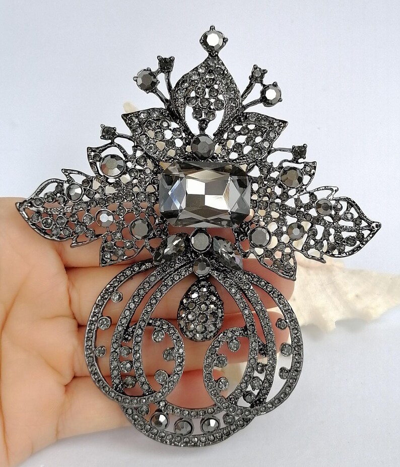 May include: A large, ornate brooch in a dark gunmetal finish, featuring a central rectangular gray crystal, surrounded by intricate filigree metalwork and numerous small, sparkling gray rhinestones arranged in floral and scroll patterns.