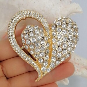 May include: A gold-coloured heart-shaped brooch, adorned with clear rhinestones. The brooch has a large heart shape with a smaller heart inside. It is held in a hand.