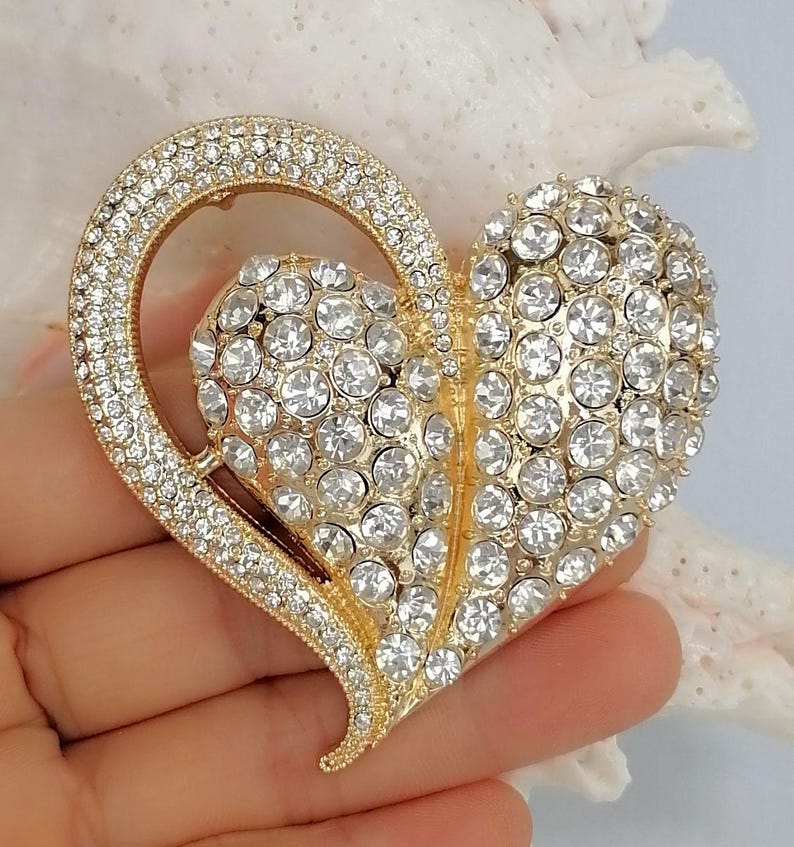 May include: A gold-coloured heart-shaped brooch, adorned with clear rhinestones. The brooch has a large heart shape with a smaller heart inside. It is held in a hand.