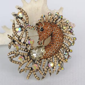 May include: A seahorse brooch with a brown body and a detailed, textured design. The seahorse is adorned with iridescent crystals and a large, clear teardrop-shaped stone. The brooch has a gold-tone metal finish and a pin closure.
