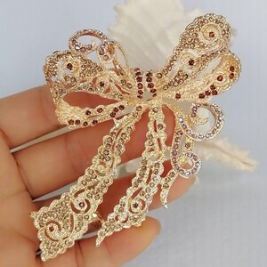 May include: A gold-toned bow brooch with intricate filigree detailing. The bow is embellished with small, sparkling crystals in shades of brown and purple. The brooch has a classic bow shape with flowing tails.