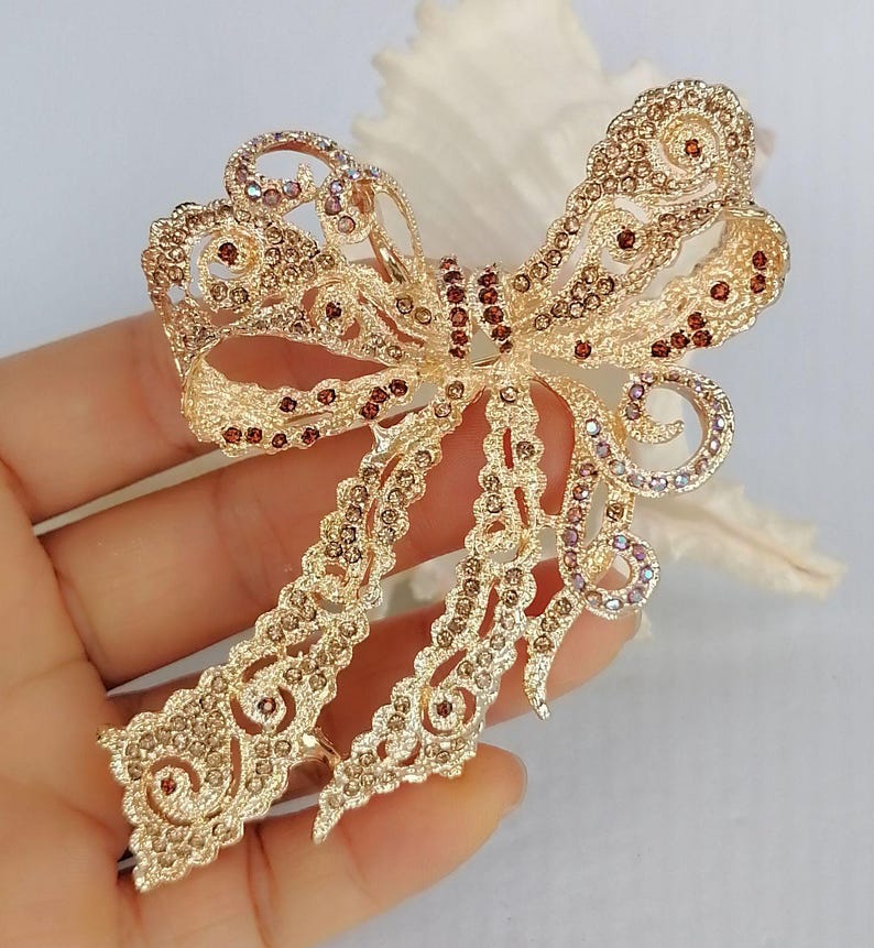 May include: A gold-toned bow brooch with intricate filigree detailing. The bow is embellished with small, sparkling crystals in shades of brown and purple. The brooch has a classic bow shape with flowing tails.